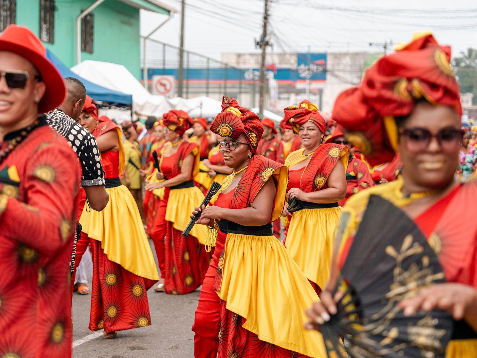 Afro-Caribbean community members in vibrant red-and-gold traditional dress and headwraps in a lively street celebration.