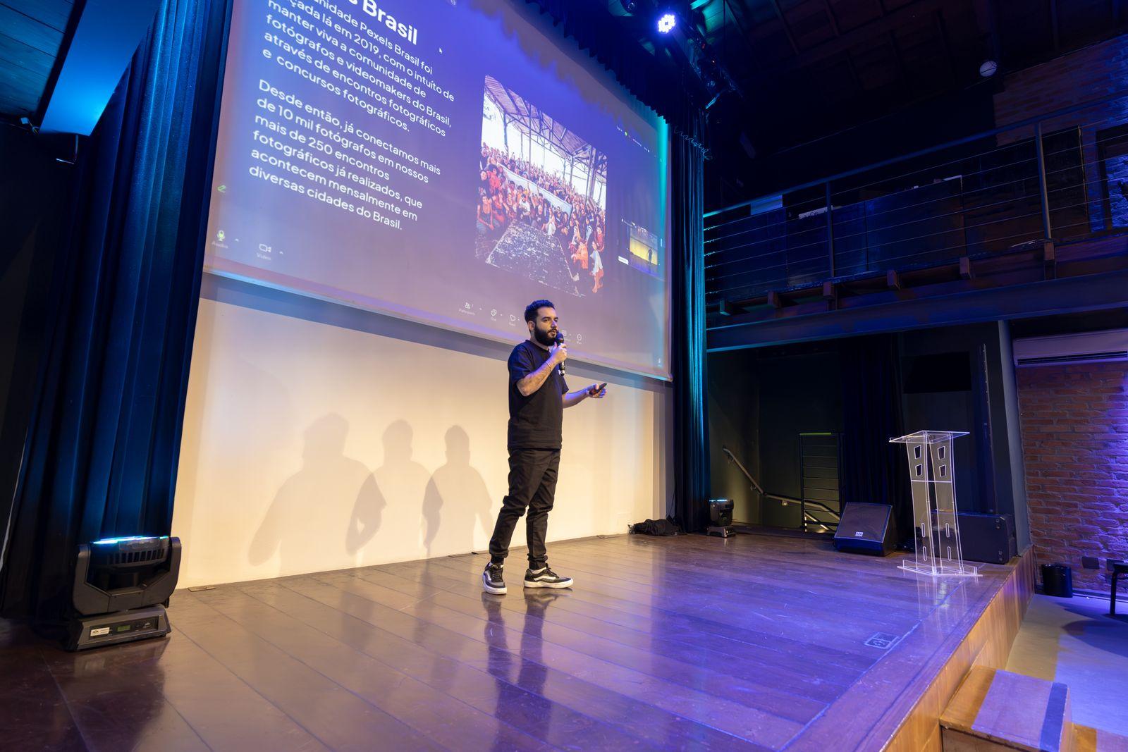Speaker presenting from a stage to a seated audience with a large presentation screen behind them