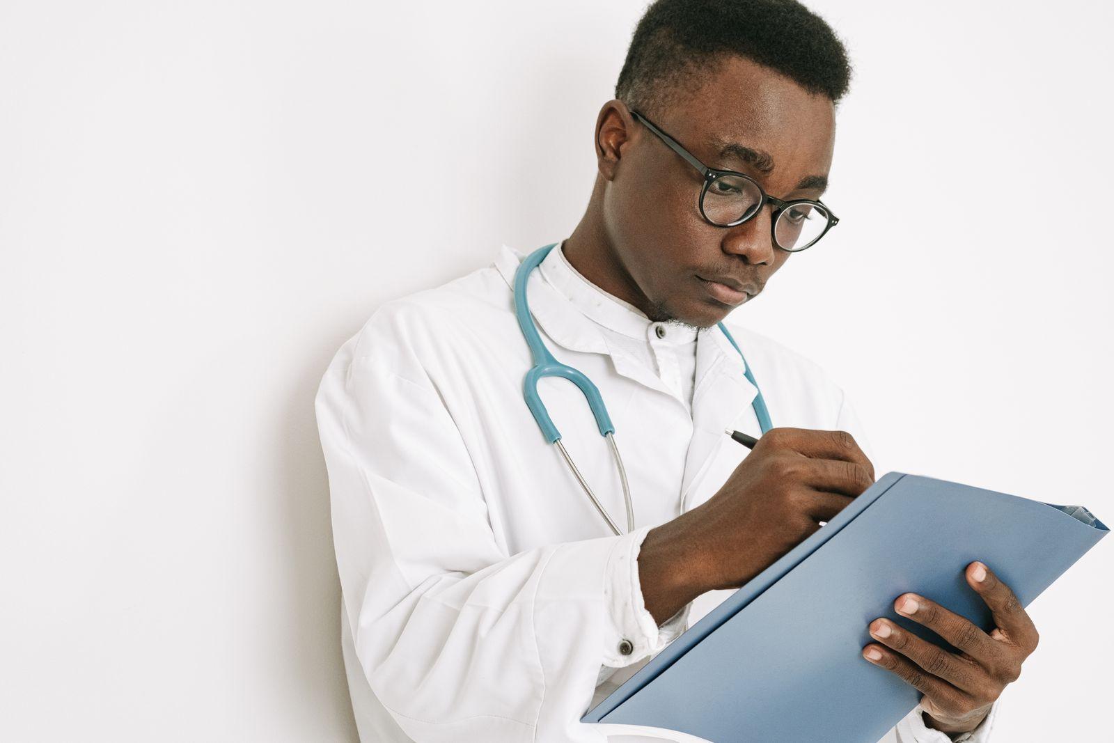 Physician in a medical gown taking notes on a patient record clipboard