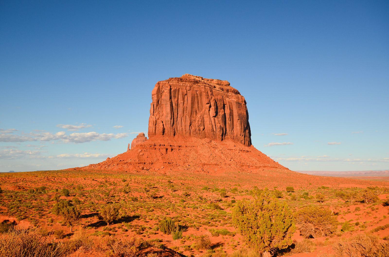 A red sandstone butte rising from the desert floor at Monument Valley, on Navajo Nation land, under a deep blue sky.
